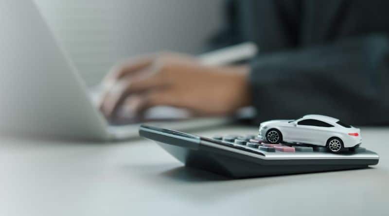 um carro de brinquedo sobre uma calculadora; ao fundo um homem digitando no notebook